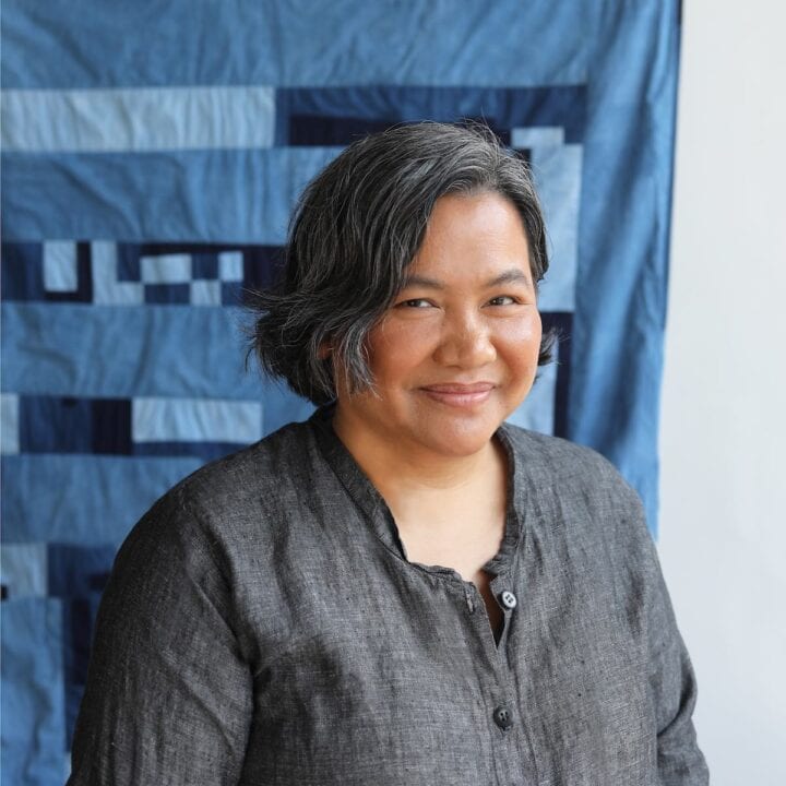 Woman with grey hair smiling in front of a blue quilt.
