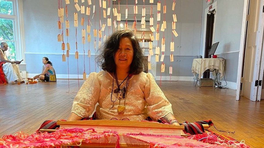 Woman weaving with pink yarn in a room.