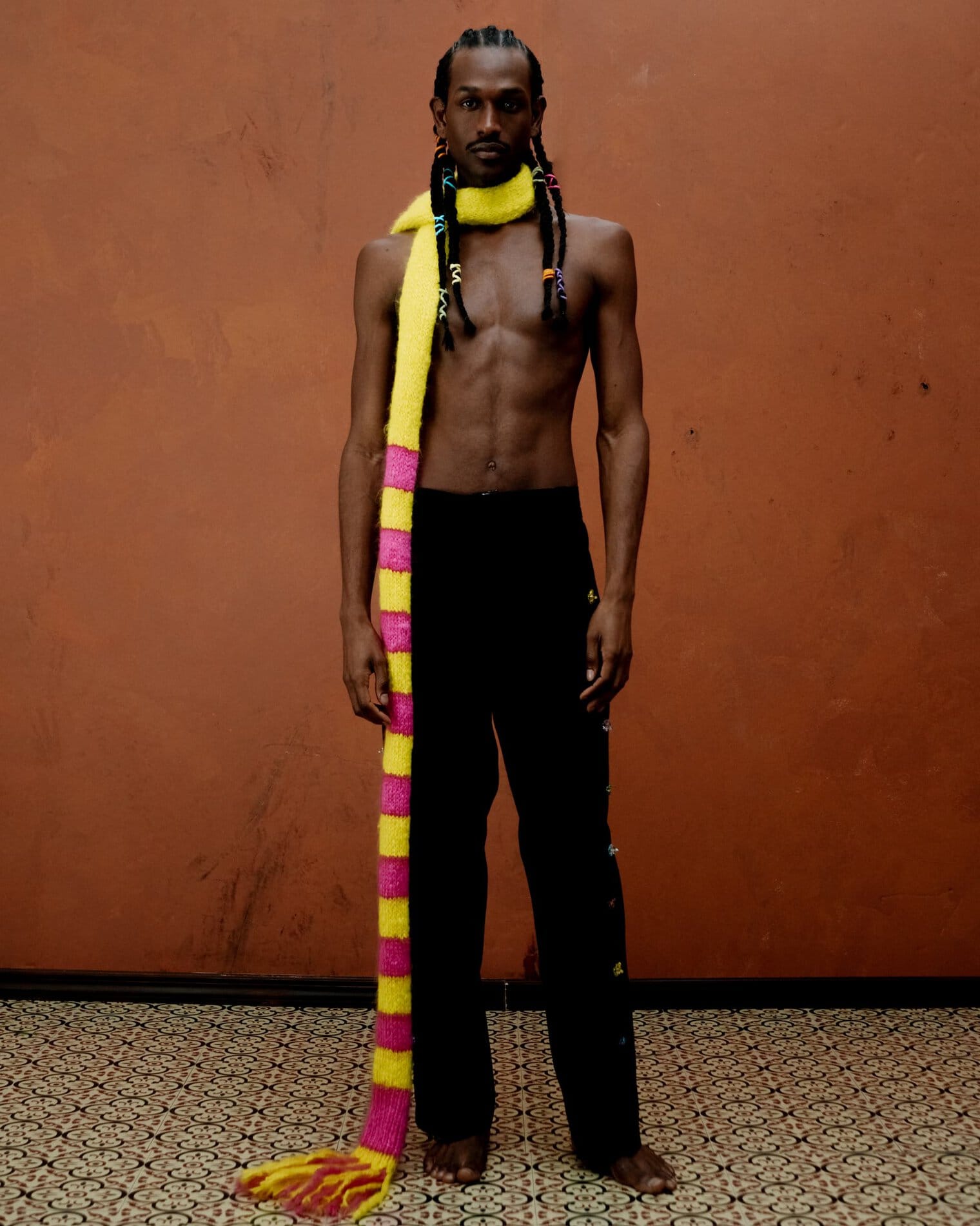 A model poses shirtless in black pants with bejewelled embellishments down the side and a long yellow and pink striped scarf.
