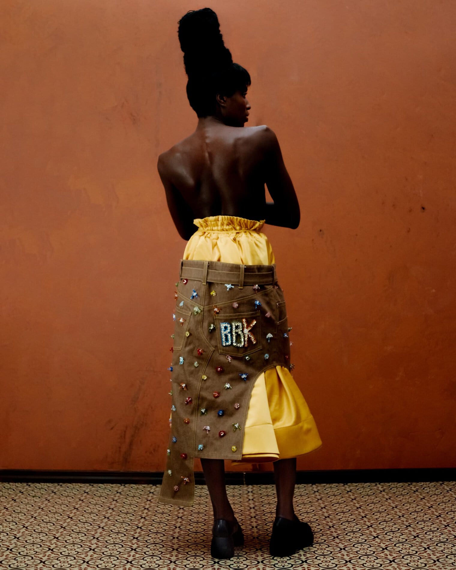 A model poses topless with her back turned to the camera, wearing a high-waisted yellow skirt underneath a belted asymmetrical dark khaki skirt with allover multicolored bejeweled embellishments and “BBK” on the right back pocket.

