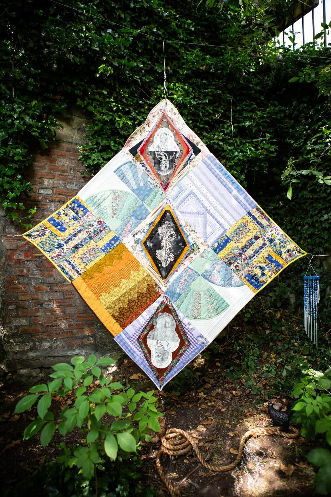 A nine-block quilt hung diagonally next to blue windchimes, surrounded by trees, with a thick rope coiled on the ground below.
