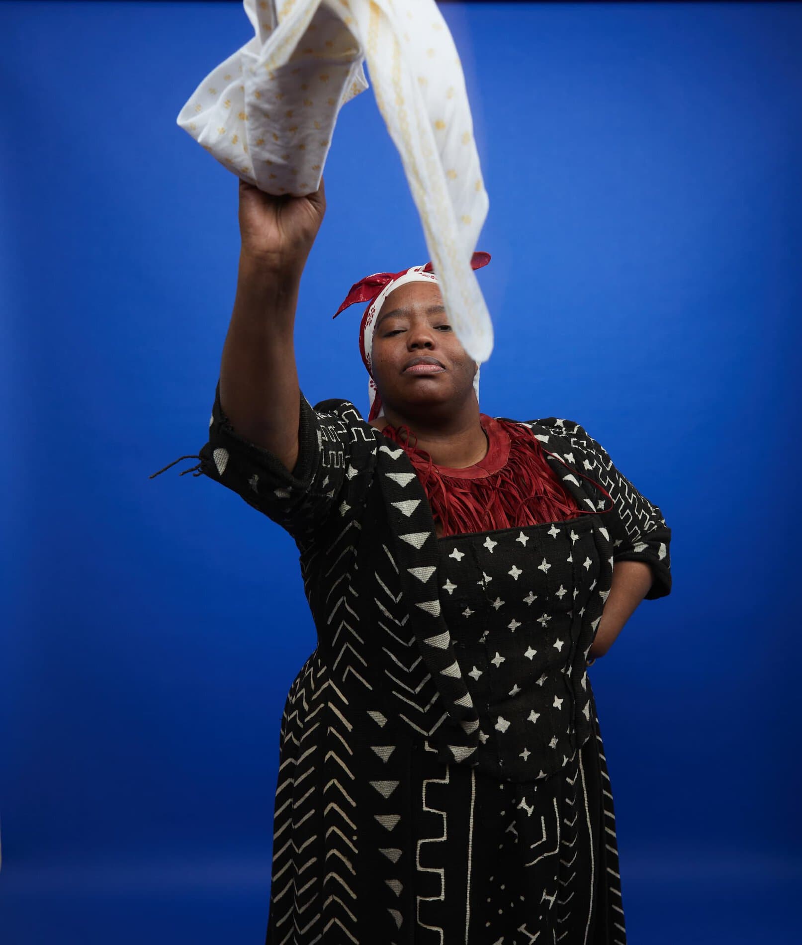 A Black woman standing in front of a blue background wearing a black and white geometric printed dress, a fringed red neck piece, and a red and white headwrap, and waving a white and yellow handkerchief.