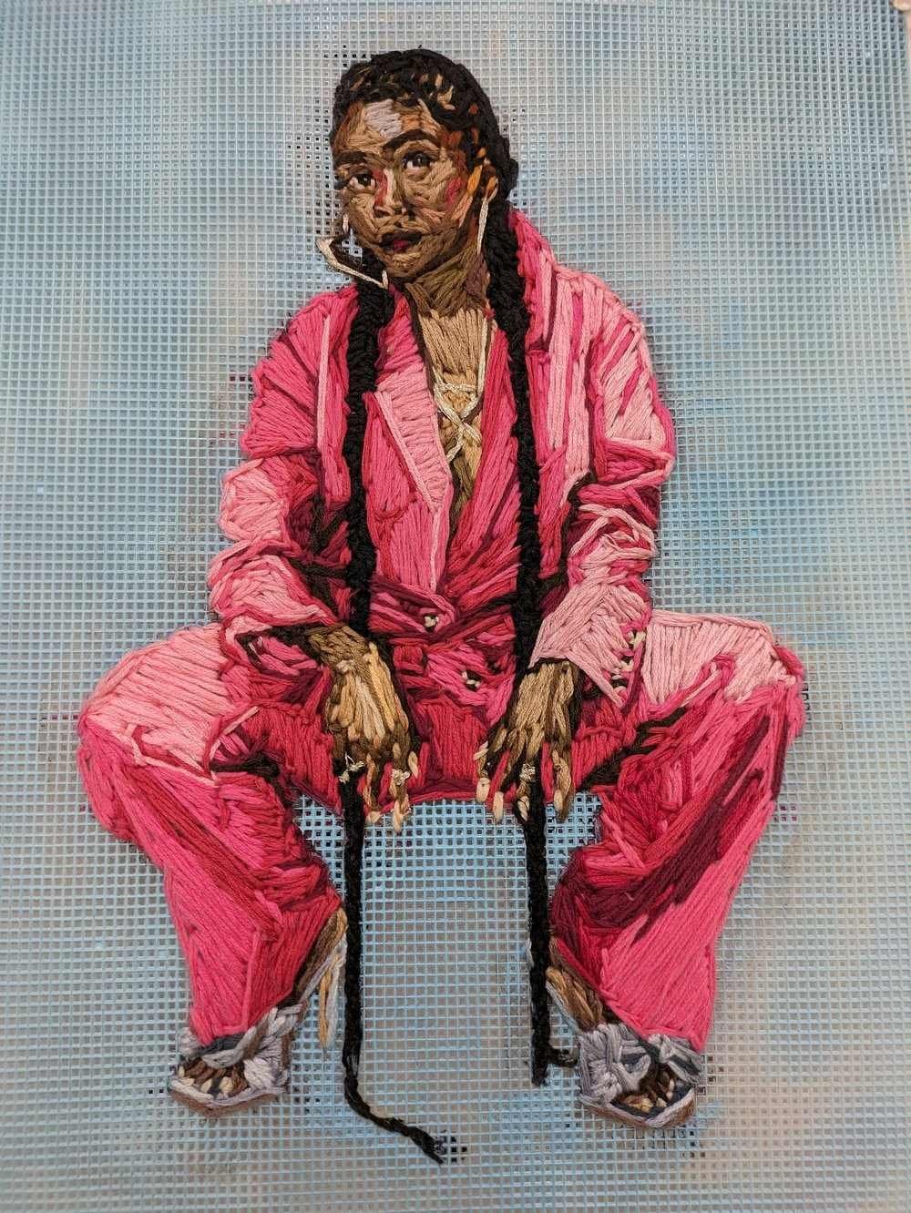 An embroidered portrait of a Black woman in a squatting position wearing large heart-shaped earrings, a pink suit, two long braids, and silver heels.