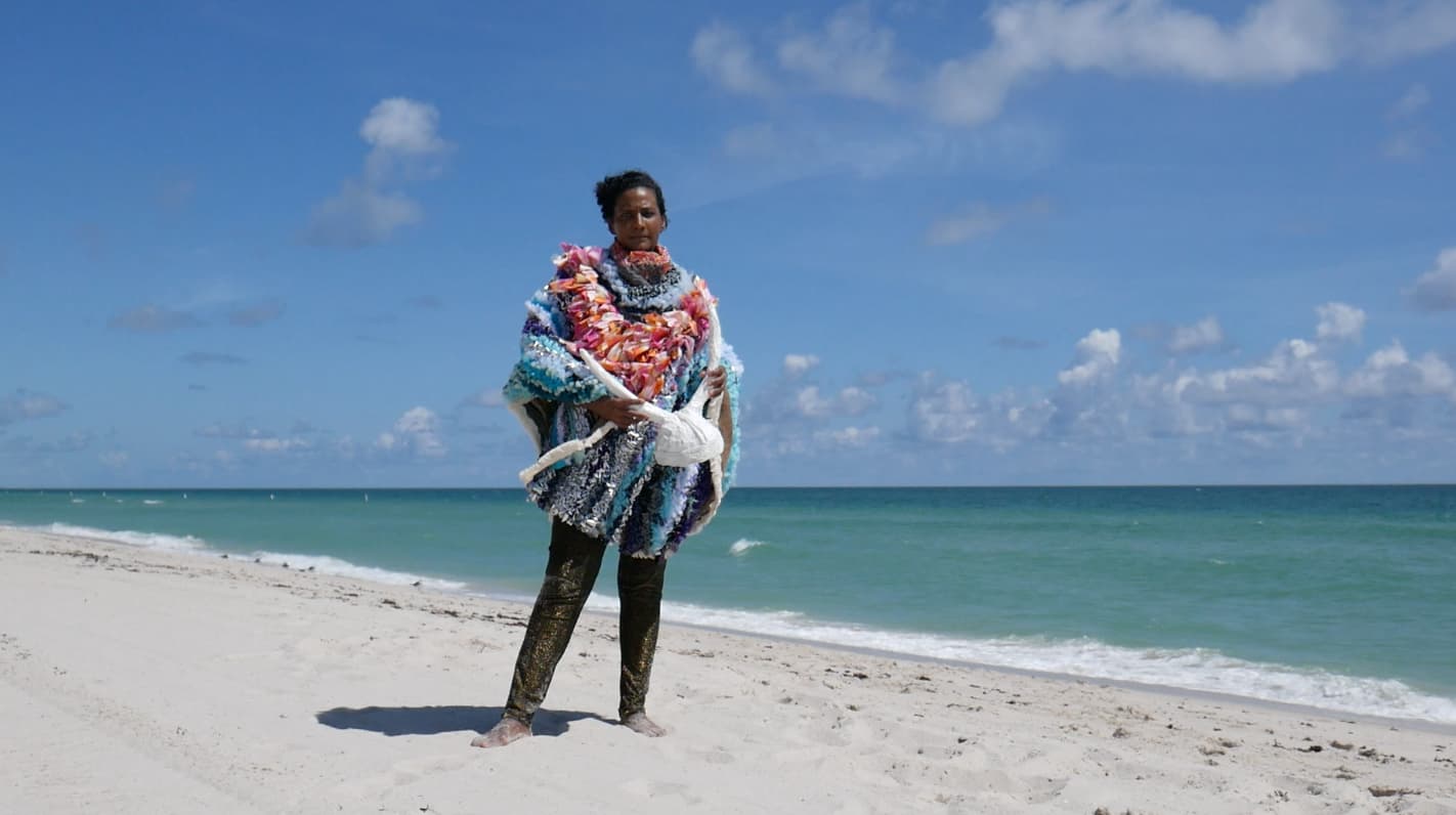 A woman standing in the sand with the ocean behind her, wearing a colorful, textured outfit and holding a white horned headpiece.
