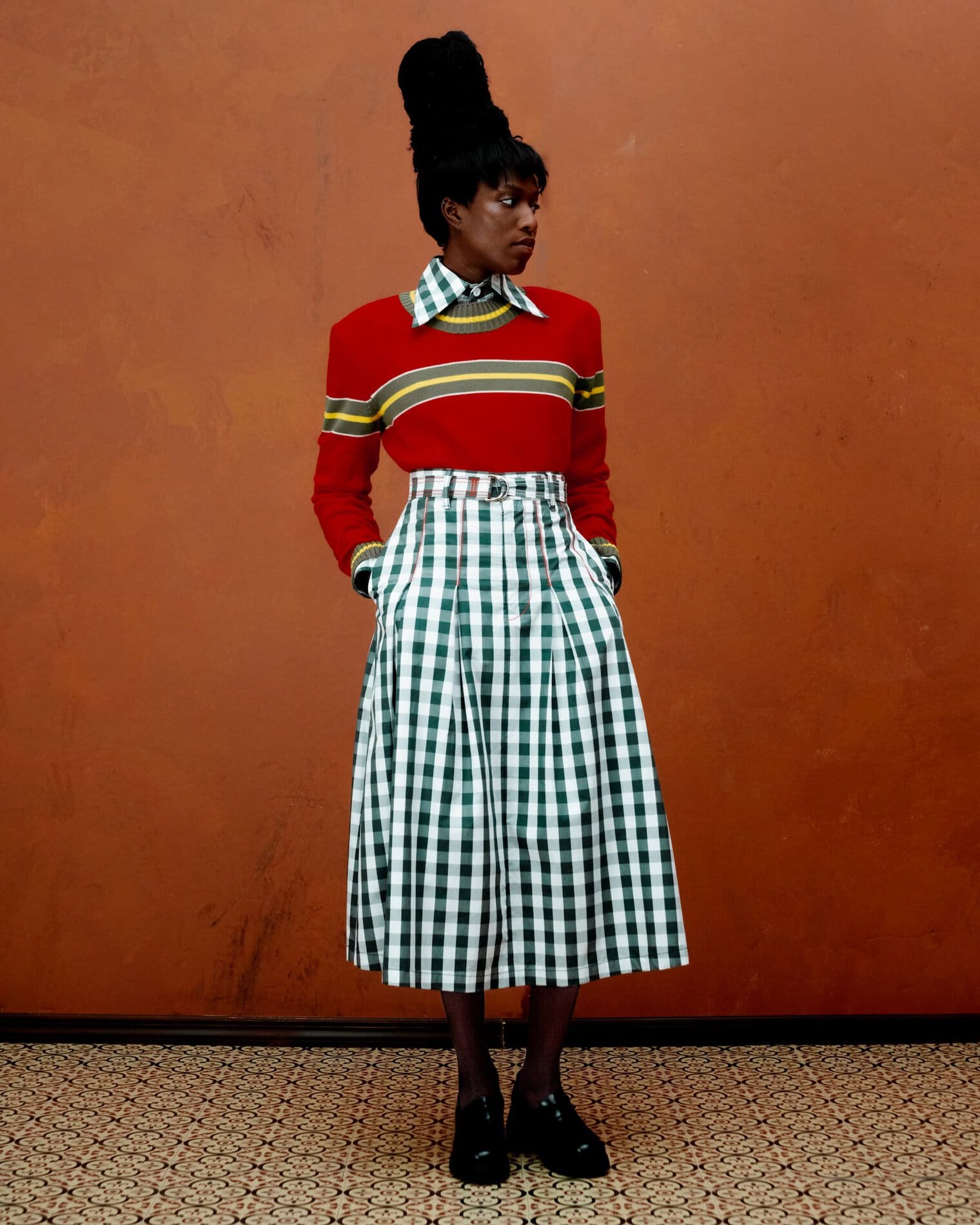 A model poses in a dark green and white gingham midi skirt, collar and sleeves, and a red sweater with green and yellow striping.
