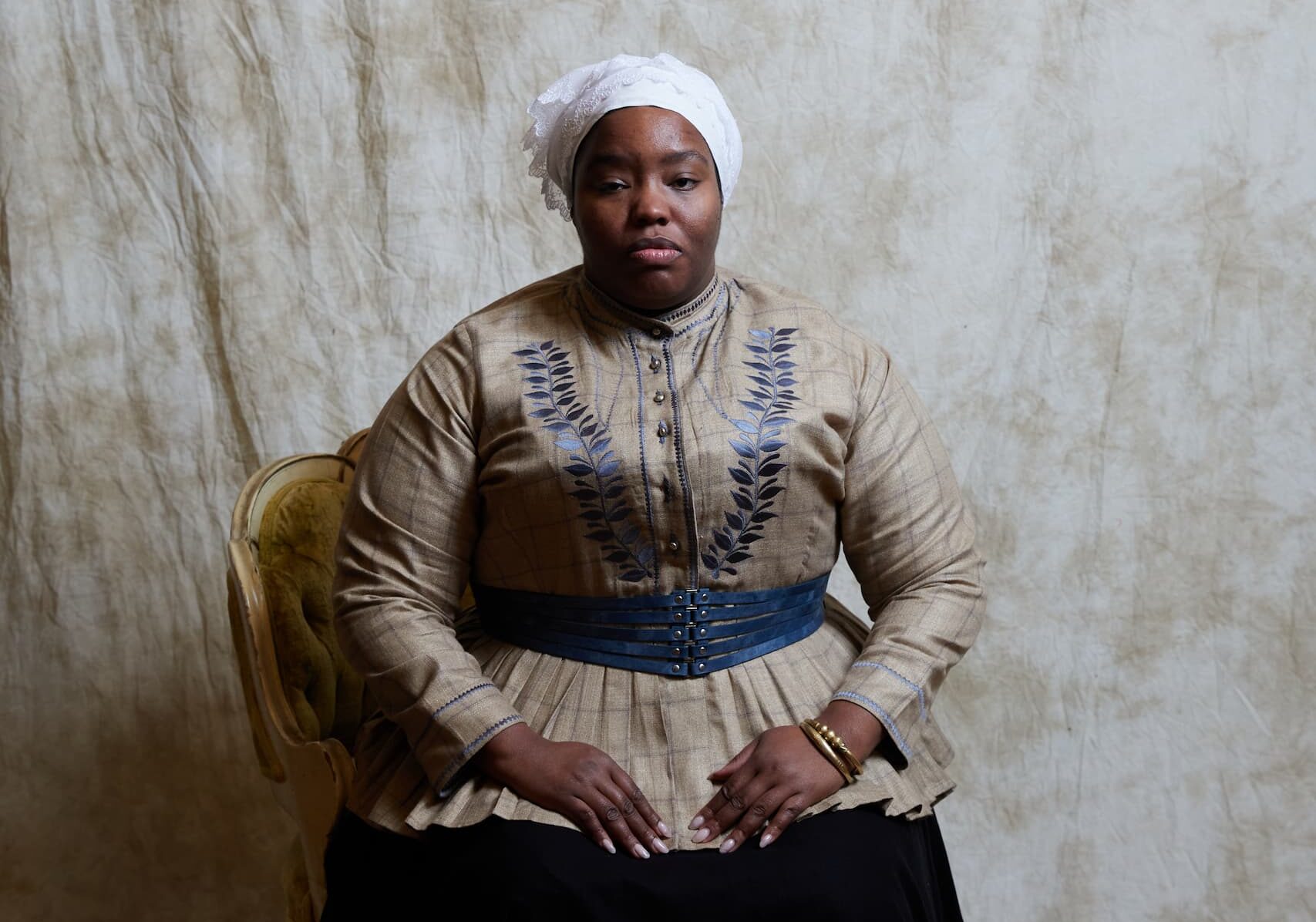 A Black woman sits with hands in lap on an upholstered chair in front of a dyed beige background wearing a white handkerchief on her head and a tan fitted jacket with dark blue embroidery across the chest and a strapped dark blue belt around her waist.
