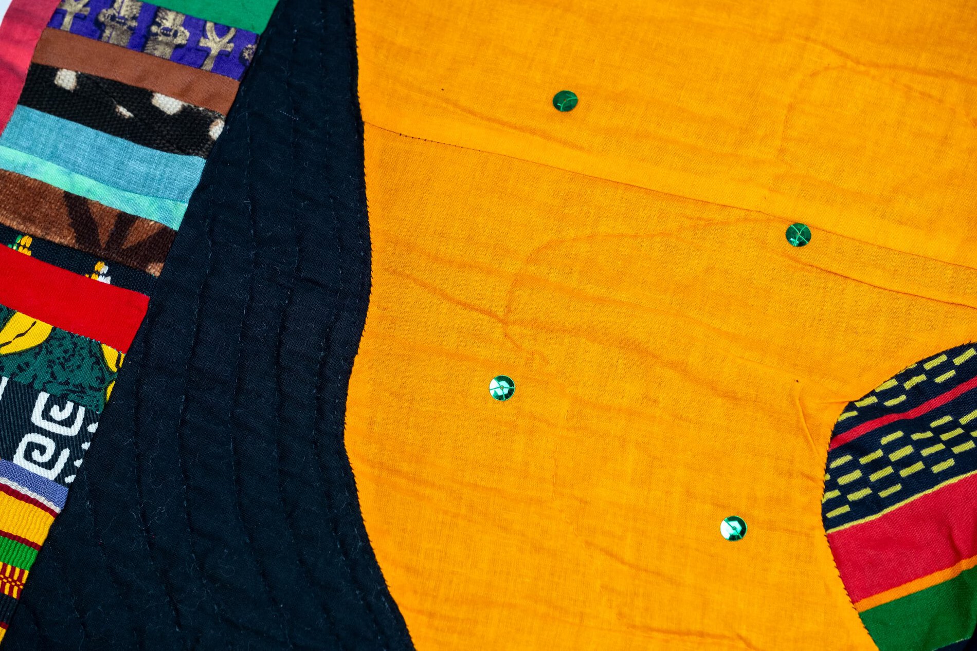 Detail of a quilt featuring many colorful patterned strips of fabric and solid black and orange shapes with sequins.