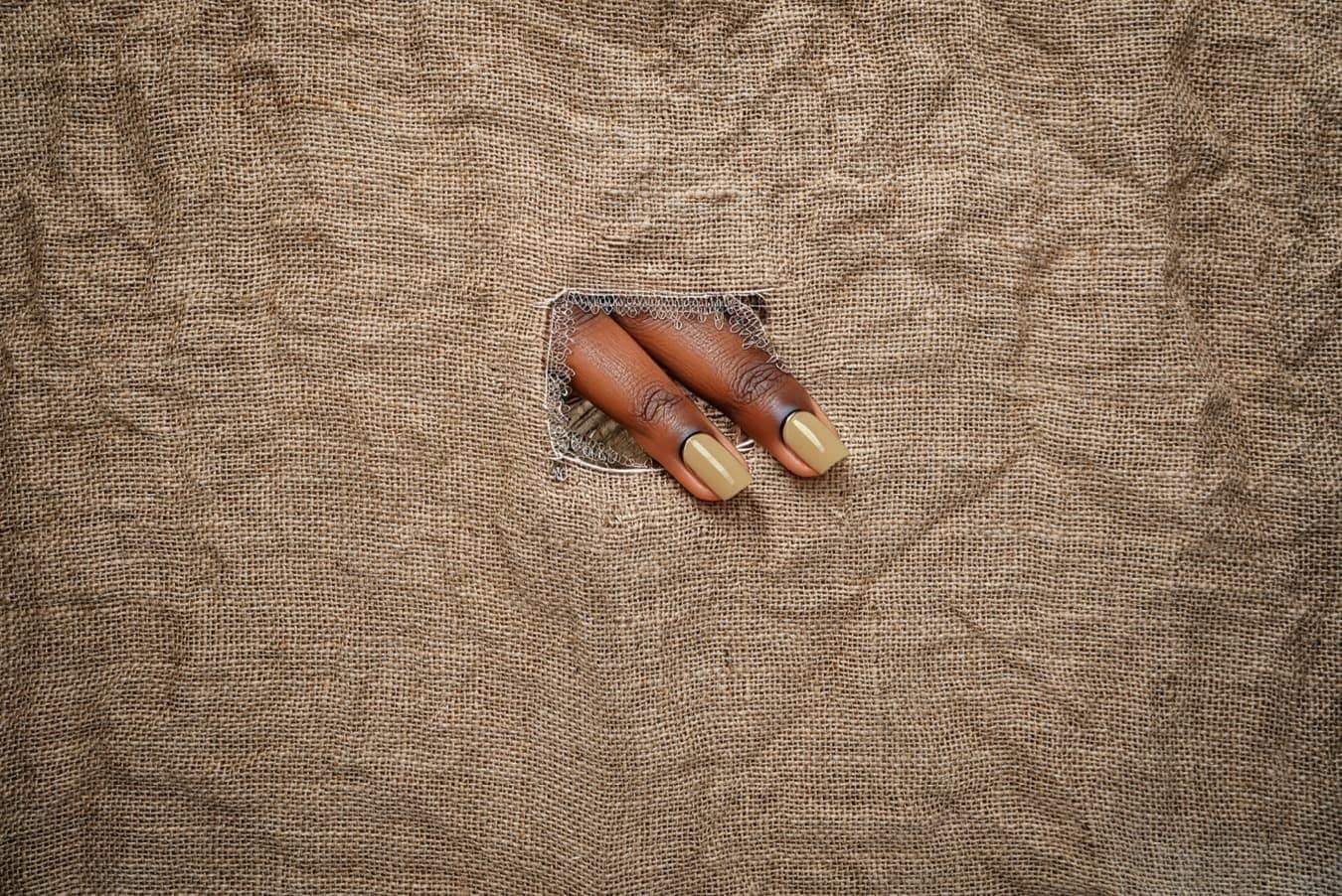 Two Black fingers poking diagonally out of a rectangular hole in a sheet of beige linen.
