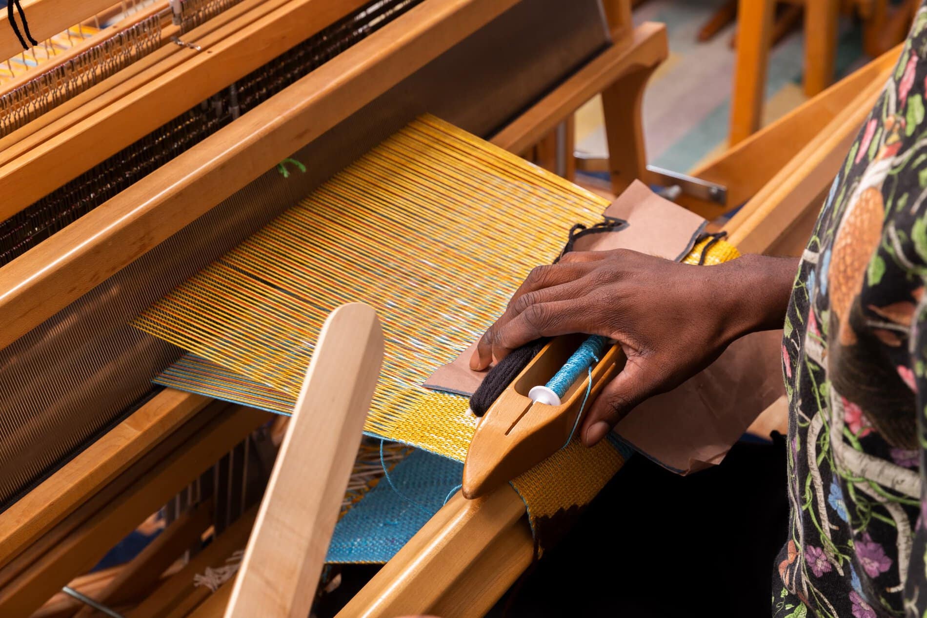 A Black hand holding a weaving shuttle in front of a standing loom with yellow warp.
