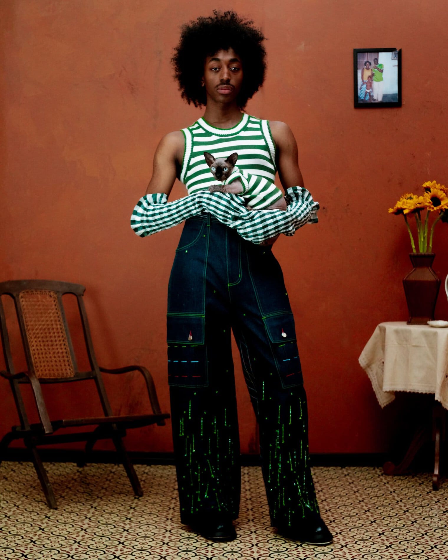 A model poses in a green and white striped tank top, green and white gingham sleeves, high waisted dark wash jeans with bright green decoration and contrast stitching, and holding a hairless cat wearing a matching top.
