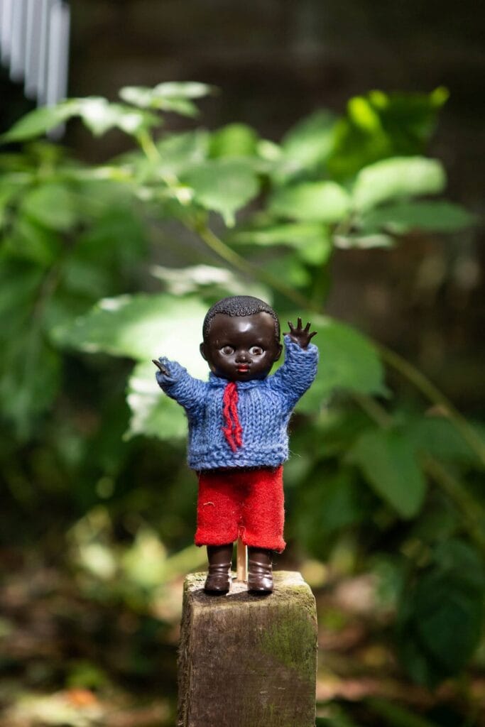 A small doll of a black child in a handknit red sweater and white skirt, standing on a wooden stump.
