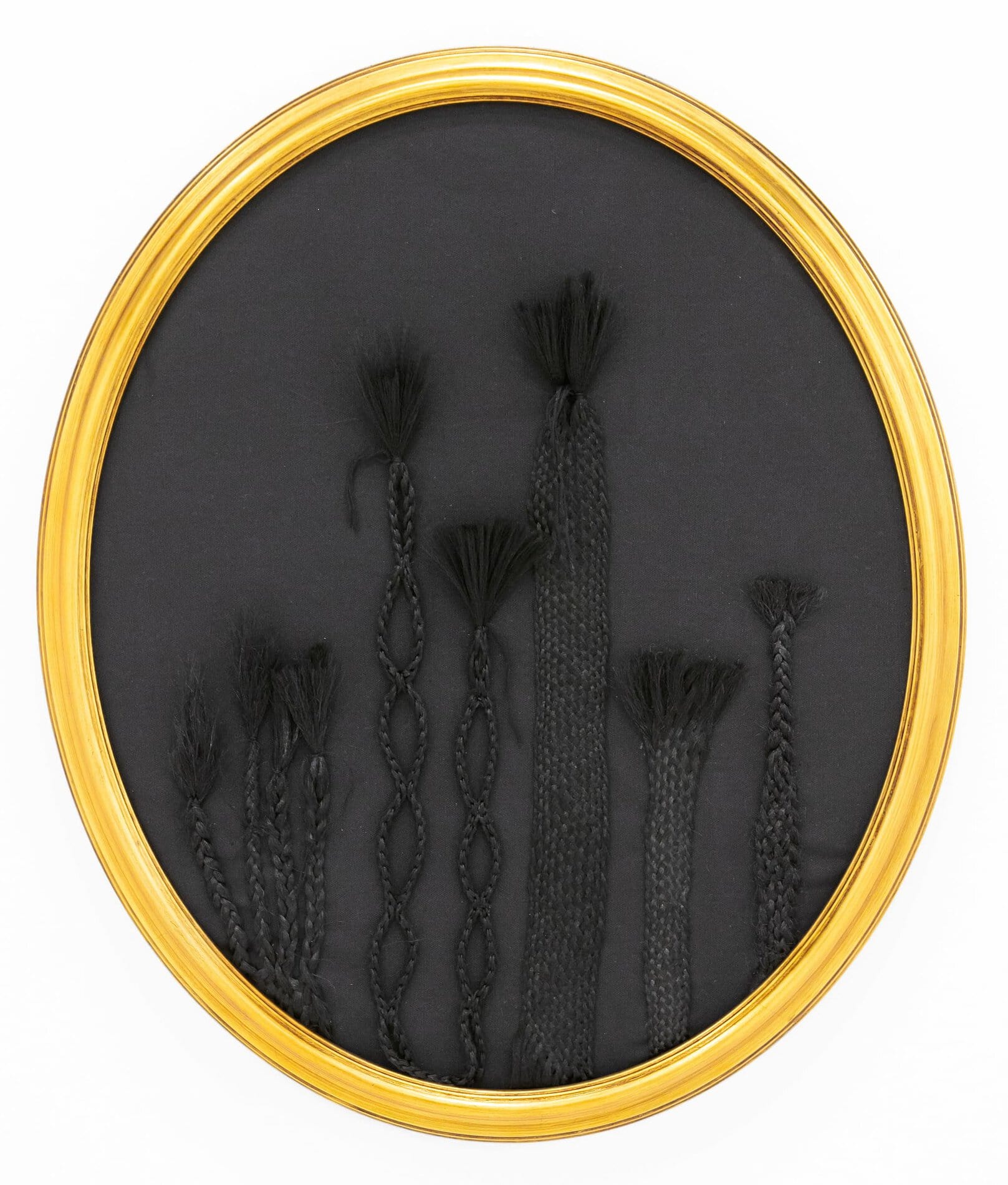 A gold oval frame containing six elaborately woven vertical plaits of black synthetic hair.
