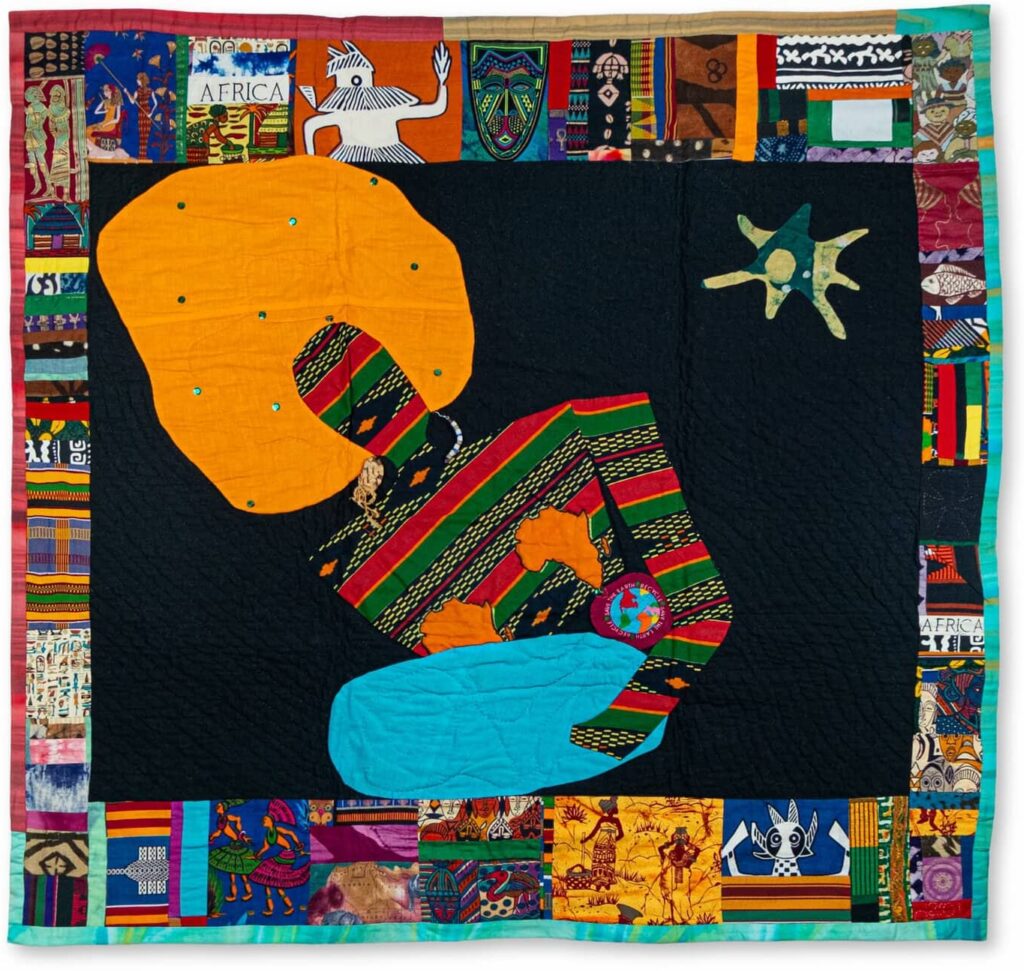 A quilt depicting a human silhouette in patterned fabric with orange hair holding a blue oval shape (front and back).
