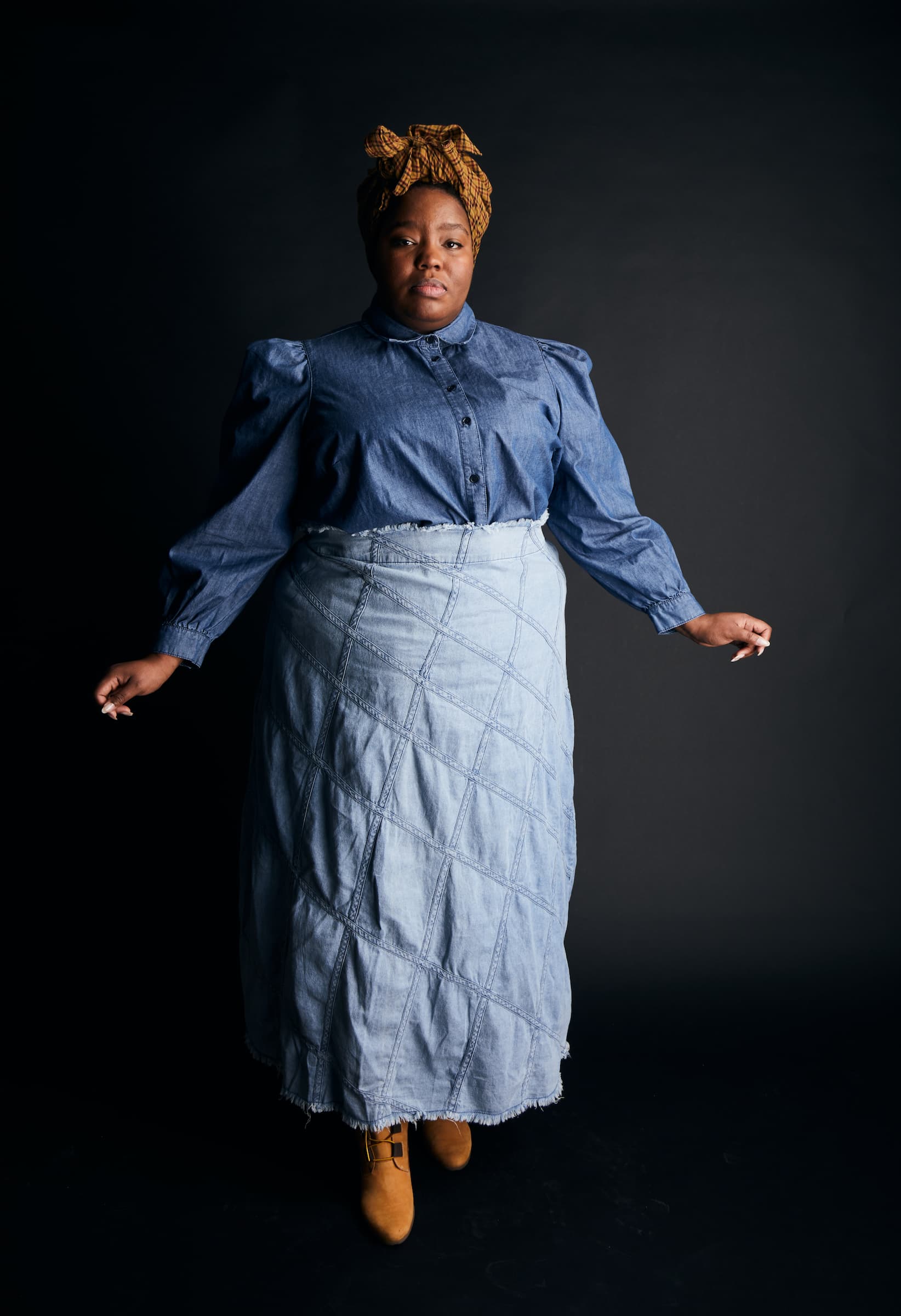 A Black woman stands in front of a black background wearing a brown headwrap, a dark denim button-down shirt, a light denim skirt, and brown boots.
