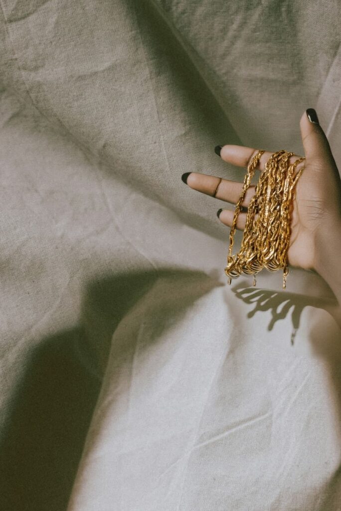 A Black hand holding gold chains above a pale sheet of fabric.
