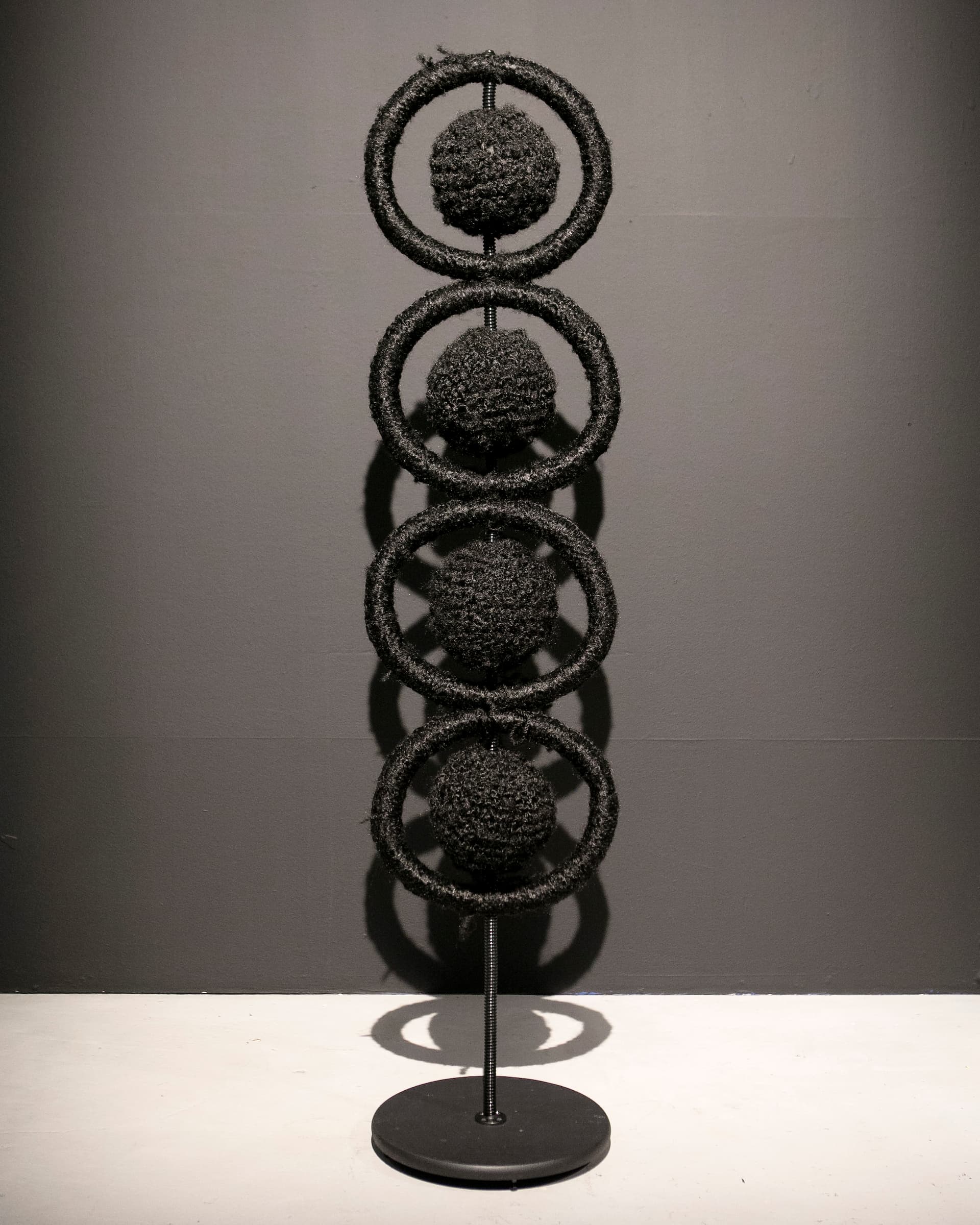 Four hollow circles wound with black synthetic hair, each encompassing a ball of black synthetic hair, stacked vertically on a stand.
