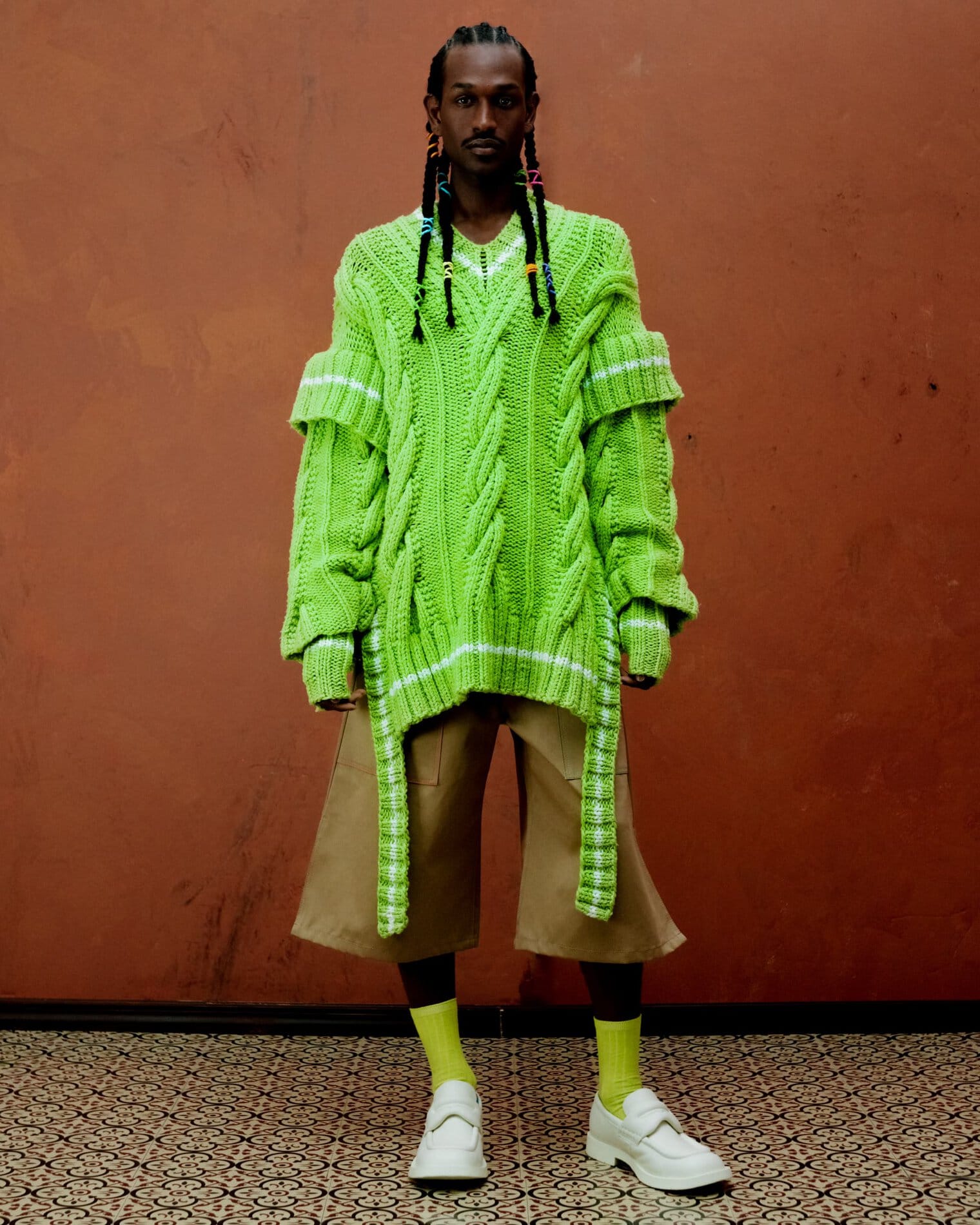 A model poses wearing a textural, oversized bright green knit sweater with white accents, flared calf-length khaki shorts, lime green socks and white loafers.
