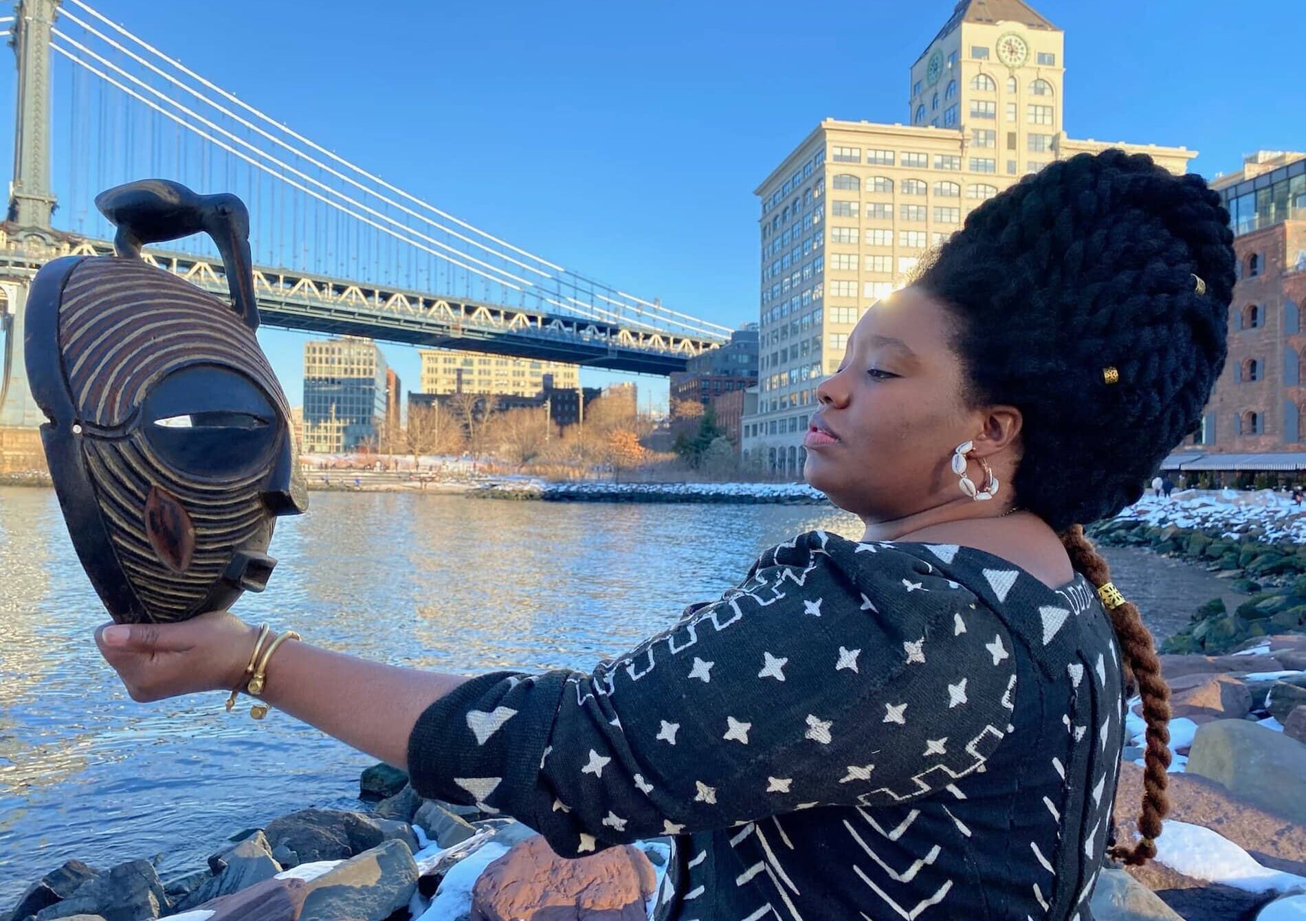 A Black woman sits on rocks near water with a building and bridge behind her, wearing a tall hairdo and braid, and holding and looking at a carved wooden mask.
