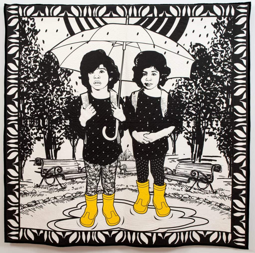 Black and white quilt of two children standing in a puddle under an umbrella, wearing yellow rainboots (front and back).

