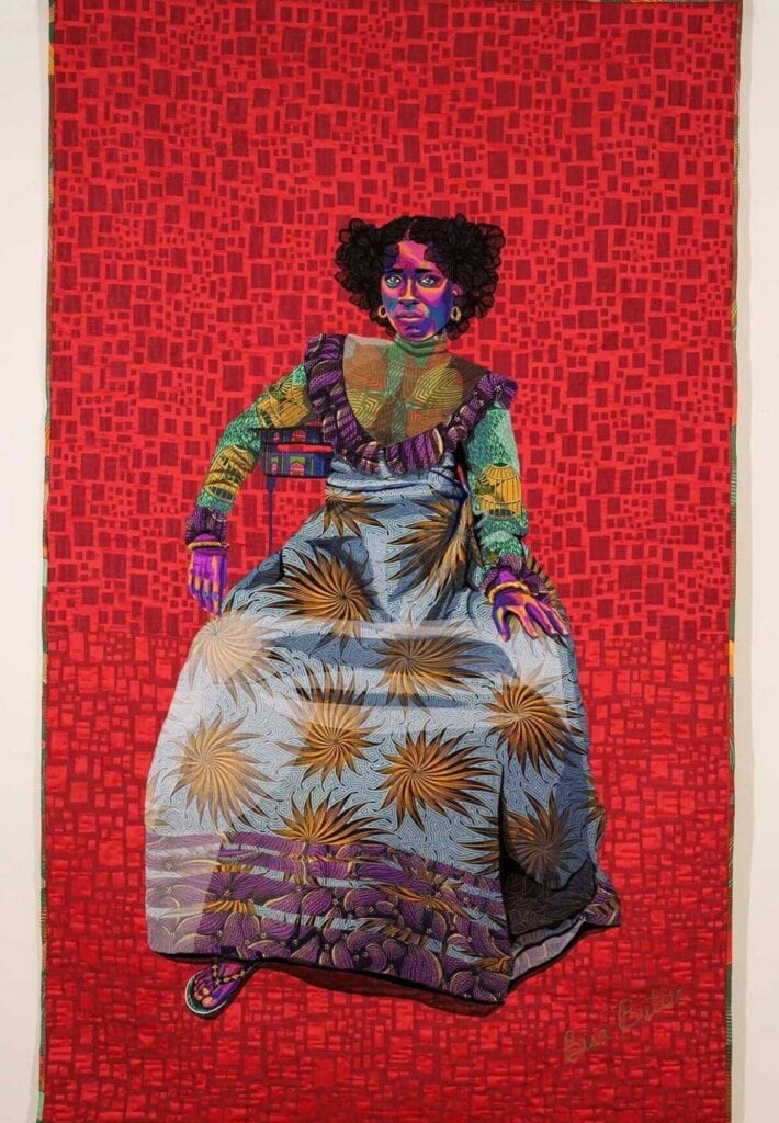 Quilted portrait of a seated Black woman in a bold printed long dress against a background of red rectangles. Bisa Butler, Asantewa.
