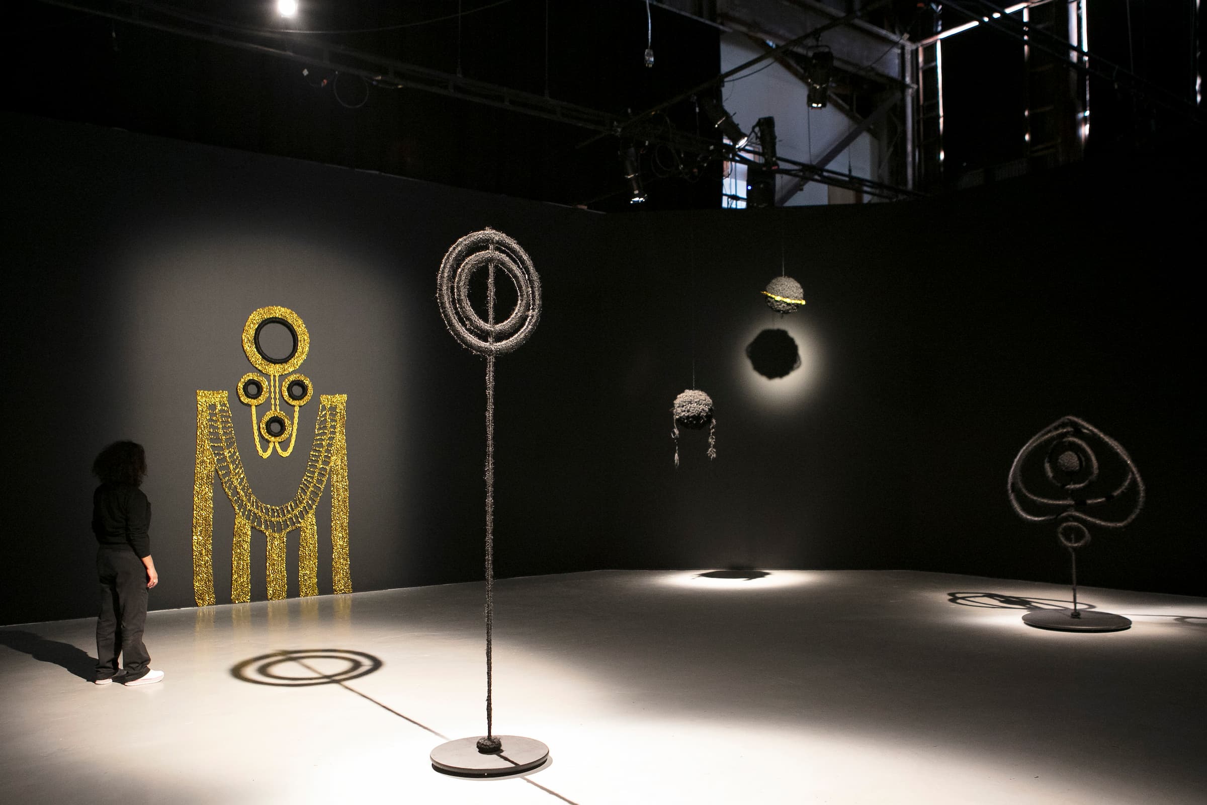 Two concentric hollow circles wound with black synthetic hair on a stand wound with black synthetic hair (foreground) in front of a gold woven wall hanging and floating black synthetic hairpieces (background).