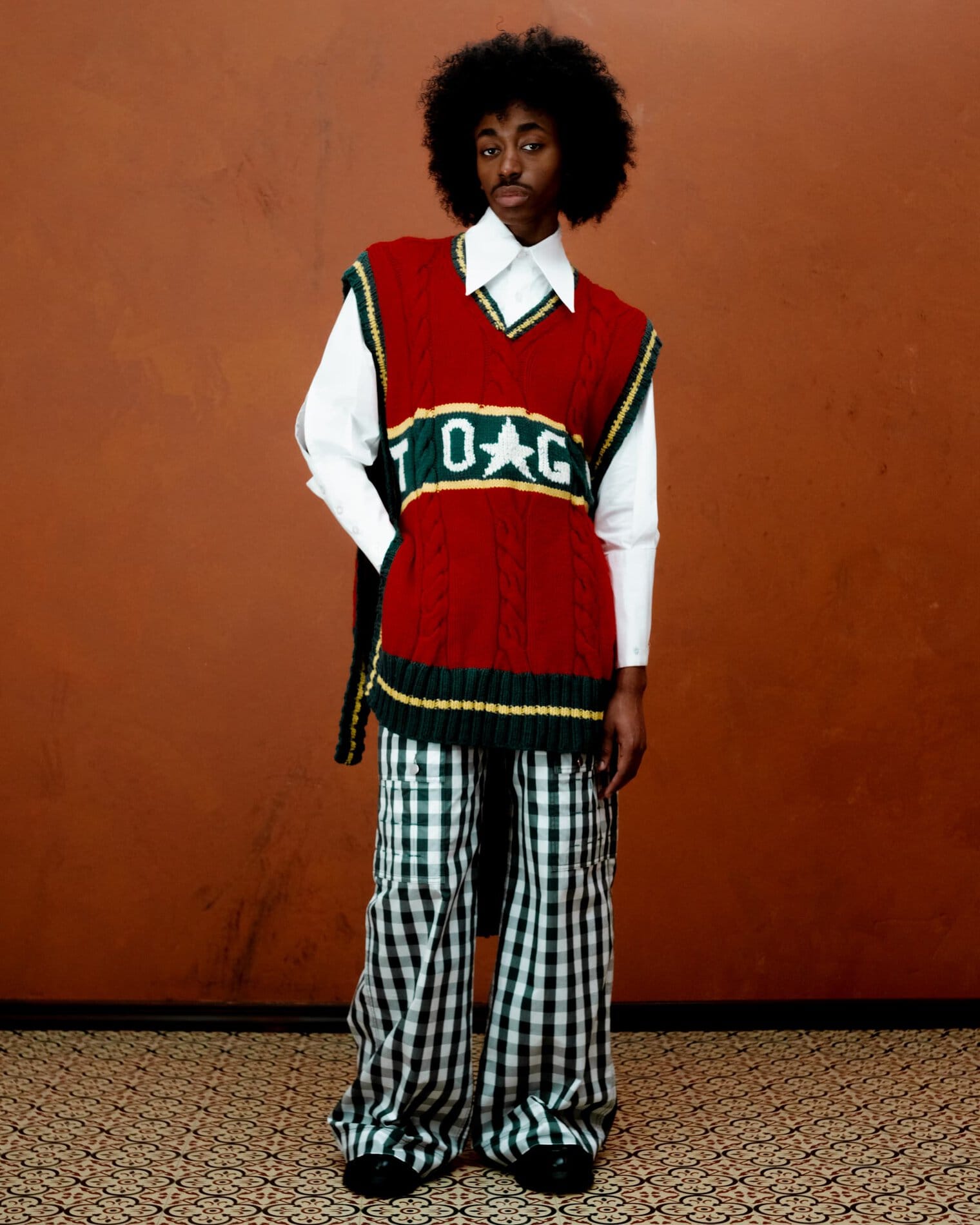 A model poses in a long sleeve white button down shirt, an oversized red sweater vest with yellow and green accents, and dark green and white gingham pants.