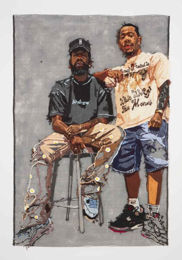 An embroidered portrait of one man sitting on a stool wearing a black hat, black t-shirt, tan pants, and white shoes, posing with another man wearing a t-shirt, jean shorts, and black sneakers.

