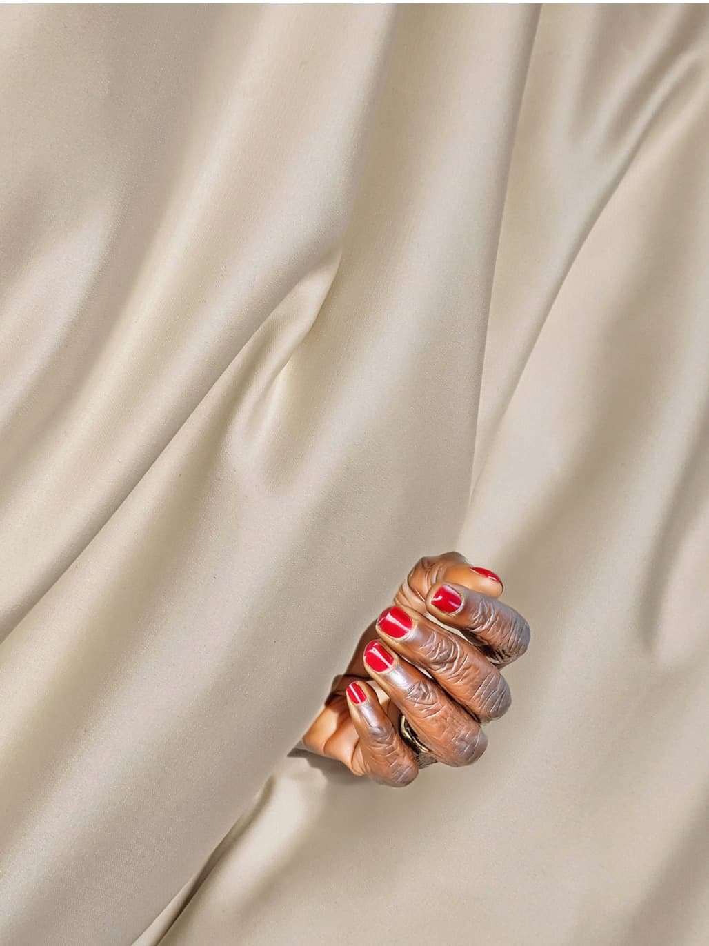 A Black hand with red painted nails peeking out from between folds of a silky white fabric.
