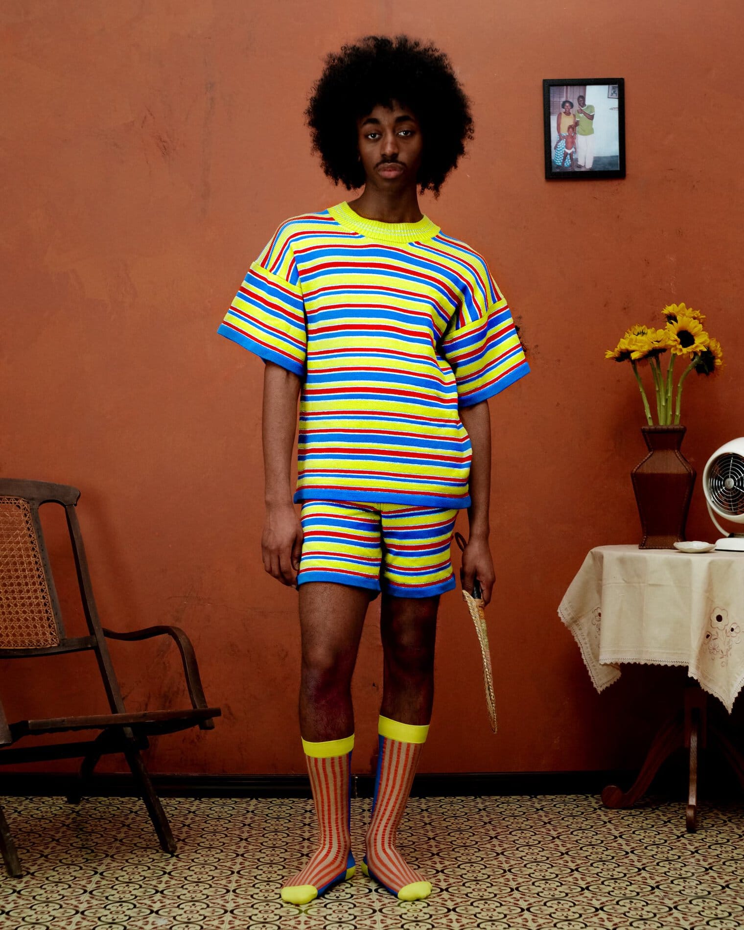 A model poses wearing a matching striped t-shirt and shorts set of yellow, blue, and red striped fabric and yellow, red and beige striped socks.

