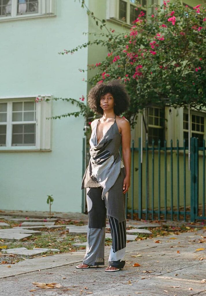 A woman in a gray and black patchworked pants and shirt set posing outside.
