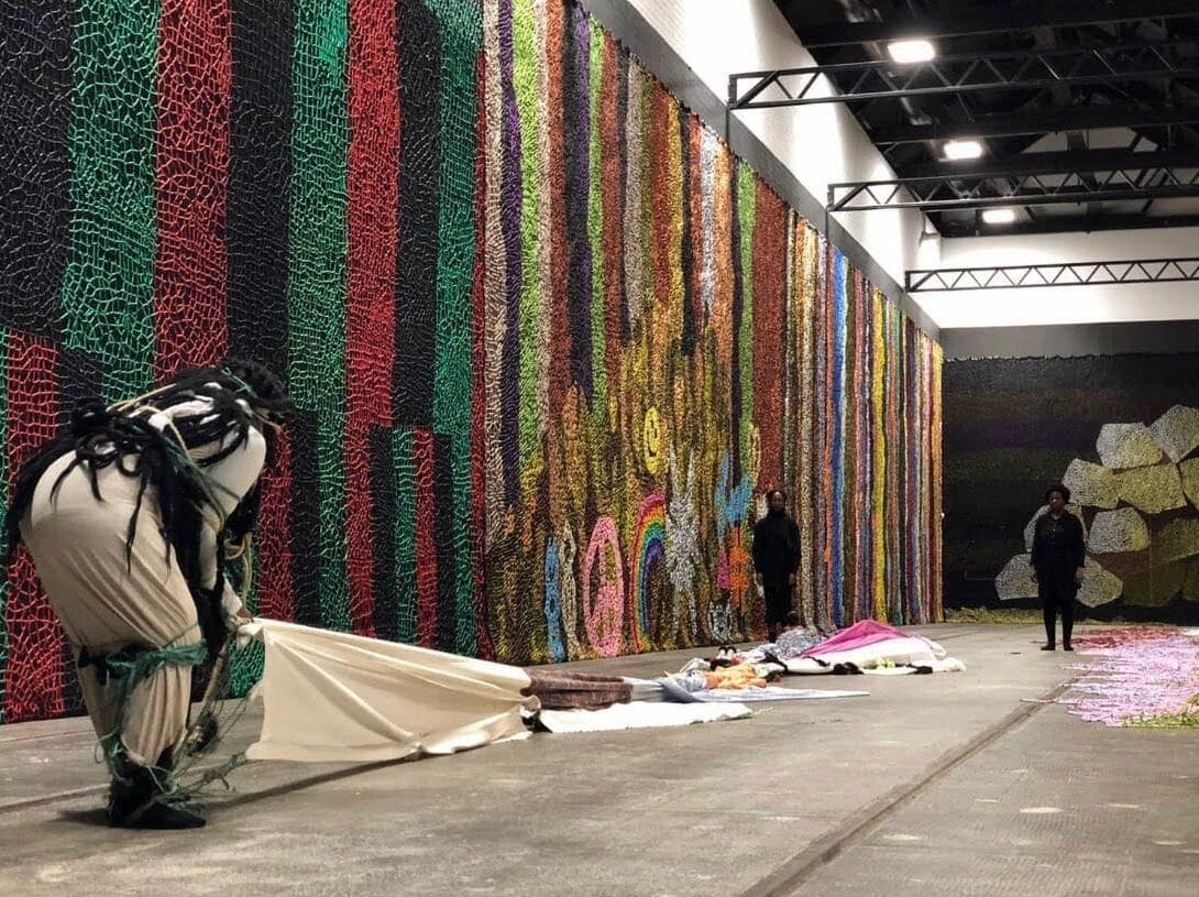 A figure standing at the end of a long train of fabrics with strips tied around her knees and feet, across from a second figure, surrounded by various large-scale fiber art pieces.
